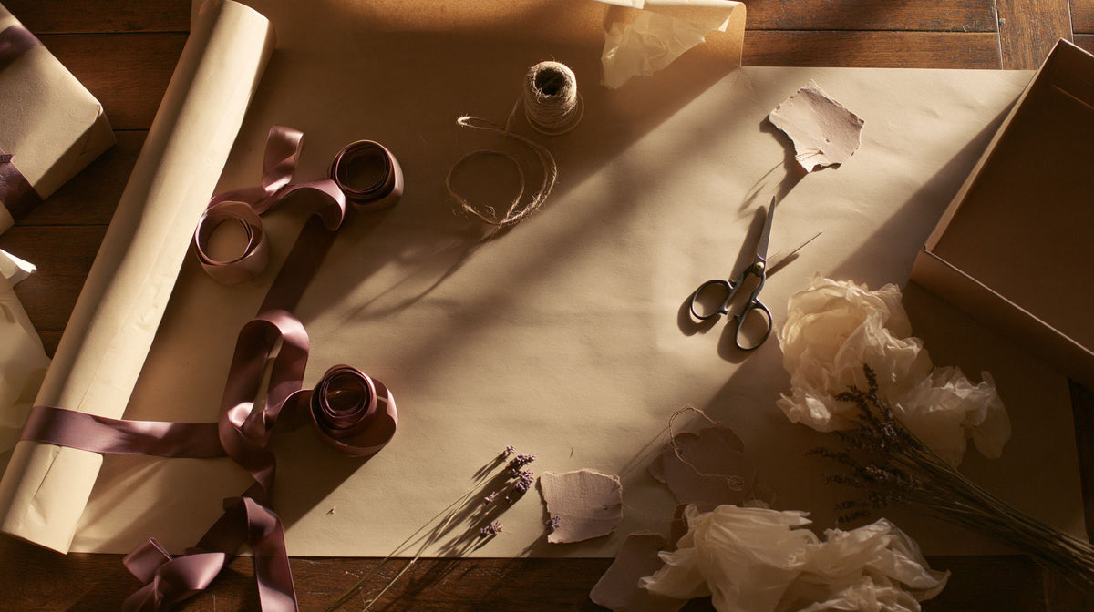 Gift wrapping in progress on a wooden table: kraft paper, plum ribbon, twine, scissors, tissue paper and lavender