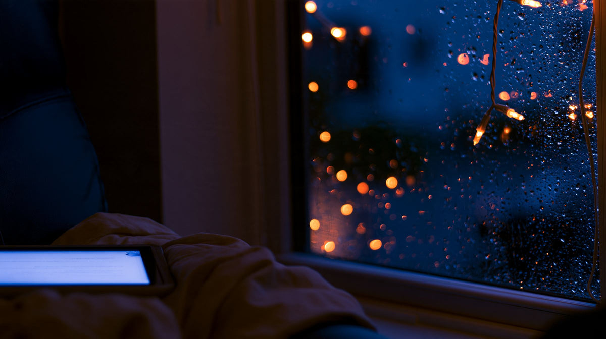 Glowing e-reader on a blanket by a rainy window at night with fairy lights reflected in the glass