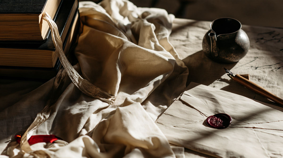 Stack of books with a ribbon bookmark beside an ink pot, a fountain pen and an envelope with a plum wax seal